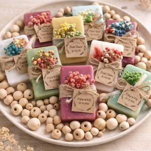 May include: A collection of rectangular soap bars in various colors, each decorated with a small bundle of dried flowers tied with twine and a brown paper tag. The tags feature printed text including Pipe & Fie and dates like 29.10.2020.