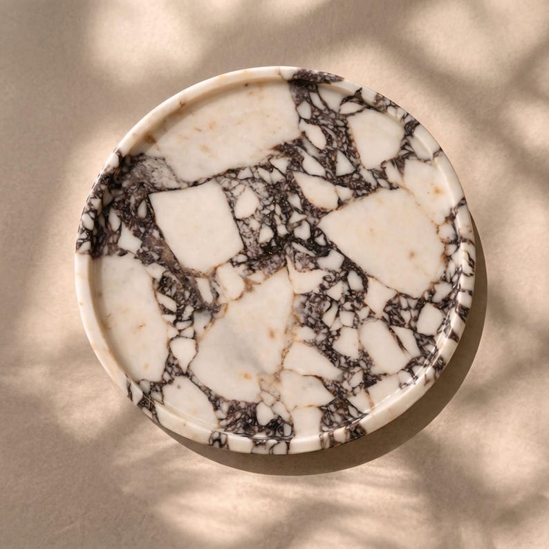 May include: Circular marble tray featuring a white and dark grey pattern. The tray has a raised edge and a smooth surface. The marble has a natural, veined appearance. The tray is set against a neutral background.