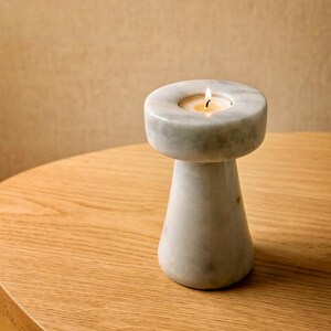 Op de afbeelding: Een marmeren kandelaar met een brandende kaars. De kandelaar is wit en grijs, met een ronde bovenkant en een conische basis. Hij staat op een houten tafel. De vlam is geel.