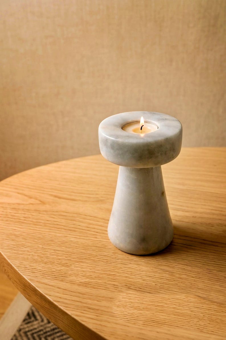 Op de afbeelding: Een marmeren kandelaar met een brandende kaars. De kandelaar is wit en grijs, met een ronde bovenkant en een conische basis. Hij staat op een houten tafel. De vlam is geel.