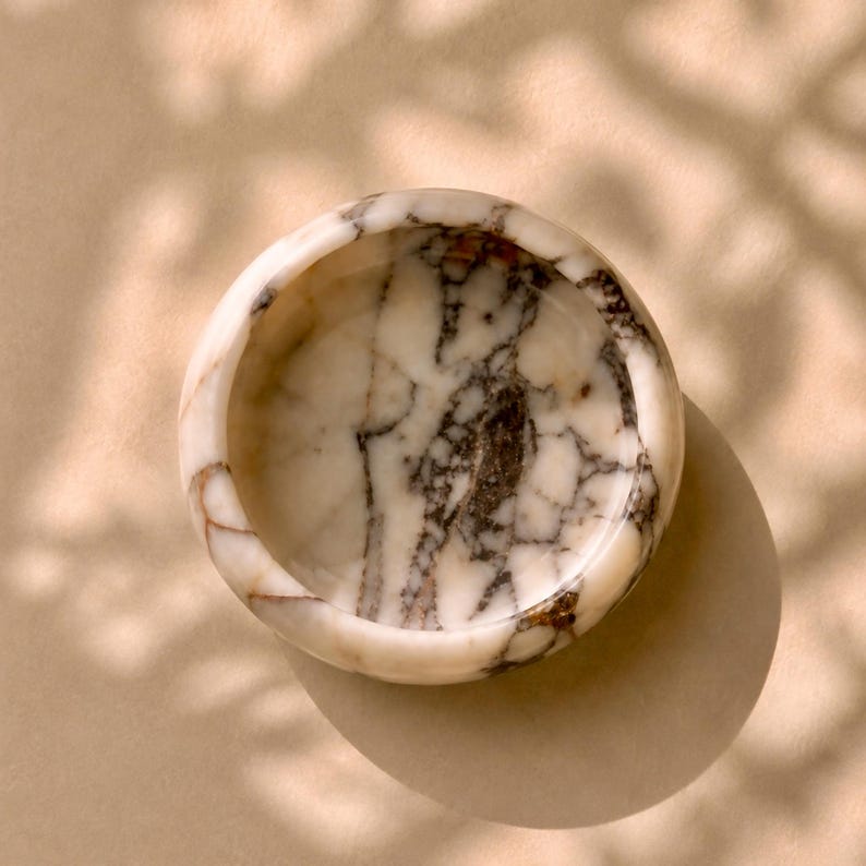 May include: A small, round marble bowl with a white and brown marbled pattern. The bowl is set against a neutral background with soft shadows, highlighting its natural stone texture and design. Ideal for small items.