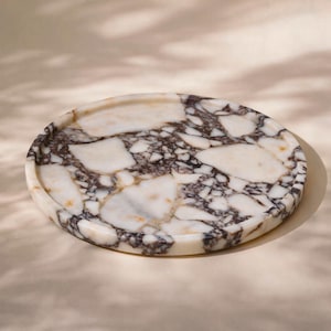 May include: A circular marble tray featuring a white and dark brown design. The tray has a raised edge and a polished surface. The marble displays a natural, veined pattern. The tray is set against a neutral backdrop.