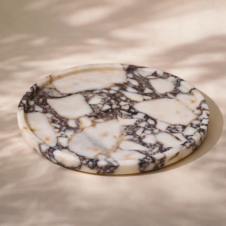 May include: A circular marble tray featuring a white and dark brown design. The tray has a raised edge and a polished surface. The marble displays a natural, veined pattern. The tray is set against a neutral backdrop.