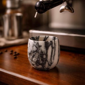 May include: A marble tumbler with a black and white pattern sits on a wooden surface. The cup is near a coffee machine, suggesting its use for espresso or other coffee drinks. Coffee beans are scattered nearby.