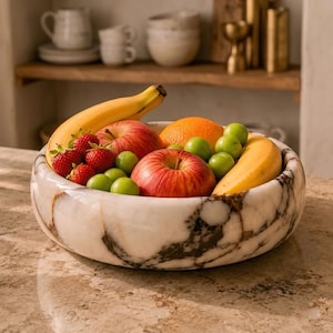 May include: A marble fruit bowl filled with fresh fruit. The bowl is white with brown veining. The fruit includes red apples, strawberries, green plums, an orange, and bananas.