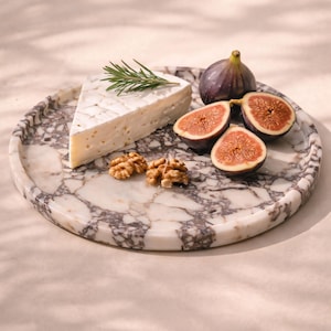 May include: A round marble serving platter with a wedge of brie cheese, fresh figs, walnuts, and a sprig of rosemary. The platter has a white and grey veined pattern. The figs are cut in half, showing their red interior.