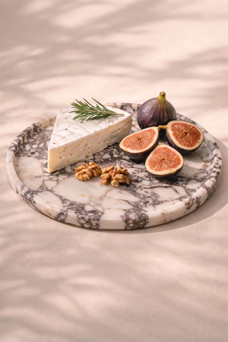 May include: A round marble serving platter with a wedge of brie cheese, fresh figs, walnuts, and a sprig of rosemary. The platter has a white and grey veined pattern. The figs are cut in half, showing their red interior.