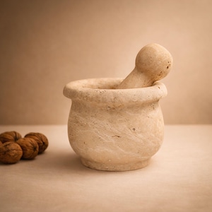 May include: A beige stone mortar and pestle. The mortar is rounded with a wide opening, and the pestle is cylindrical with a rounded top. Several walnuts are visible to the left of the mortar. The set is made of a textured stone material.