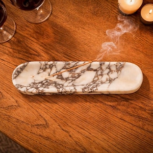 以下が含まれることがあります： 茶色と白の模様の大理石の香立て。 燃えているお香がホルダーに置かれ、煙が立ち上っています。 ホルダーは木製の表面にあり、背景にはろうそくの火とワイングラスがあります。