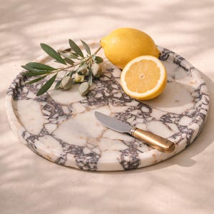 May include: A round marble serving tray with a grey and white pattern, holding a halved lemon, a whole lemon, a sprig of olives, and a small knife with a gold handle. The tray is approximately 30 cm in diameter.