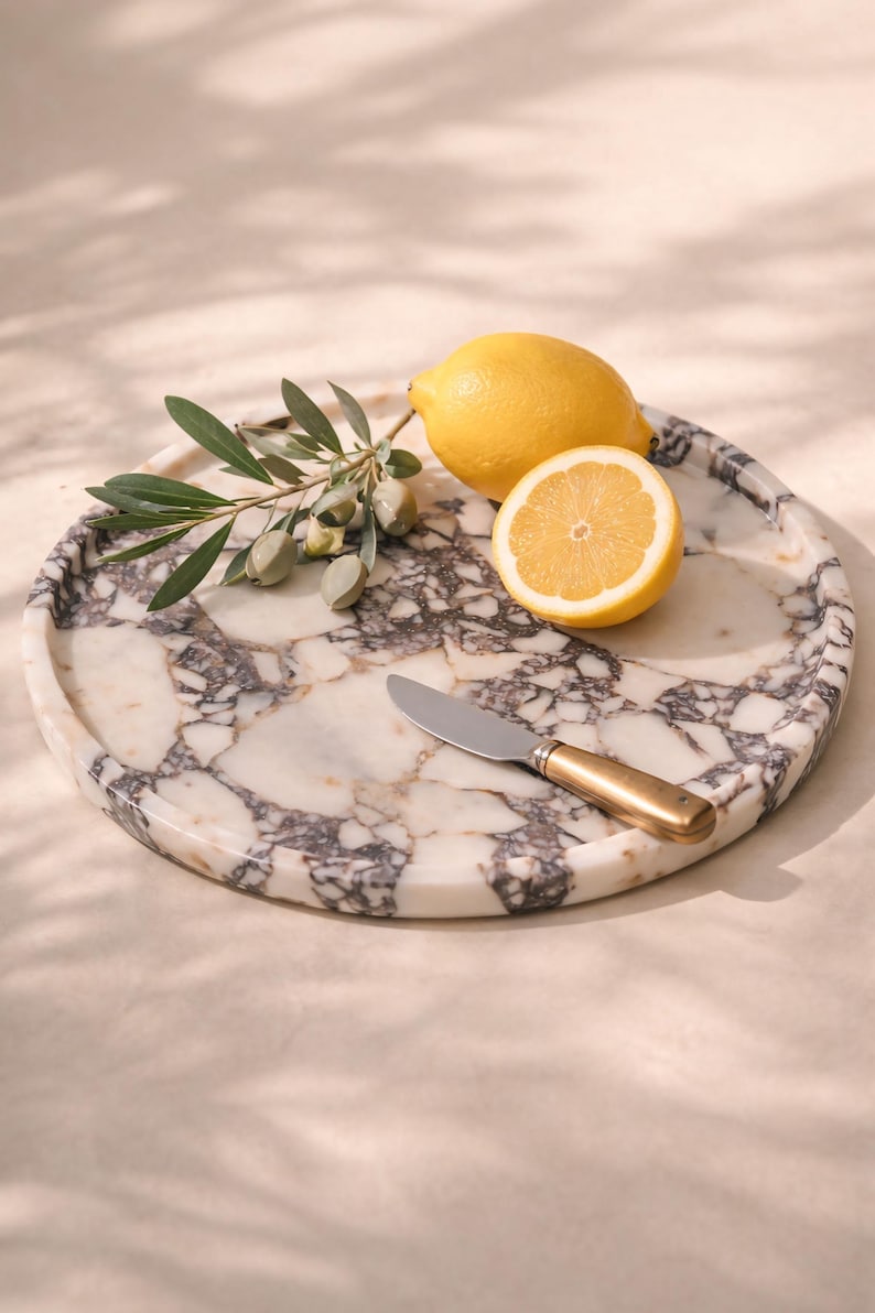 May include: A round marble serving tray with a grey and white pattern, holding a halved lemon, a whole lemon, a sprig of olives, and a small knife with a gold handle. The tray is approximately 30 cm in diameter.