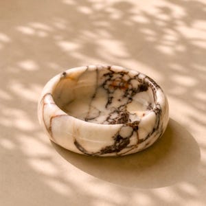 May include: A small, round marble bowl with a white base and brown veining. The bowl has a smooth, polished surface and is set against a neutral background. The bowl's design is simple and elegant, suitable for holding small items.