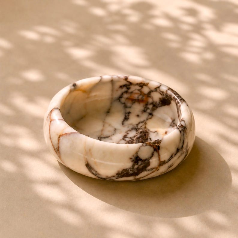 May include: A small, round marble bowl with a white base and brown veining. The bowl has a smooth, polished surface and is set against a neutral background. The bowl's design is simple and elegant, suitable for holding small items.