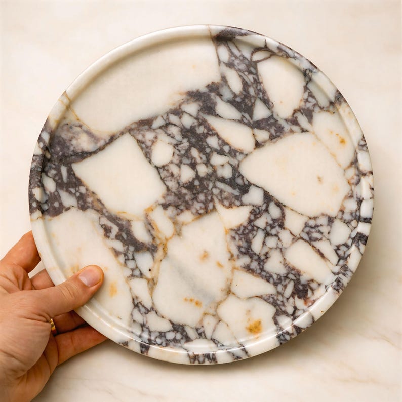 May include: A circular marble tray featuring a white and dark purple pattern. The tray is held by a hand and has a raised edge. The marble has a natural, veined appearance, with areas of white and purple.