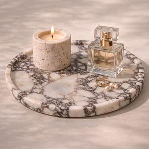May include: A circular marble tray featuring a lit candle, perfume bottle, ring, and pearls. The tray displays a white and grey veined pattern. The candle is in a stone holder. The perfume bottle is square with a gold top.