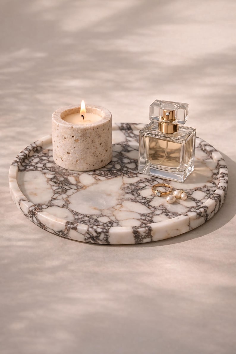 May include: A circular marble tray featuring a lit candle, perfume bottle, ring, and pearls. The tray displays a white and grey veined pattern. The candle is in a stone holder. The perfume bottle is square with a gold top.