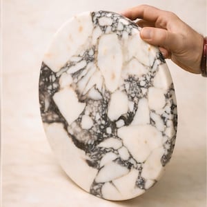 May include: An oval marble chopping board with a distinctive pattern of white, black, and tan veining. The board is held by a hand, highlighting its polished surface and natural stone variations. A decorative kitchen item.