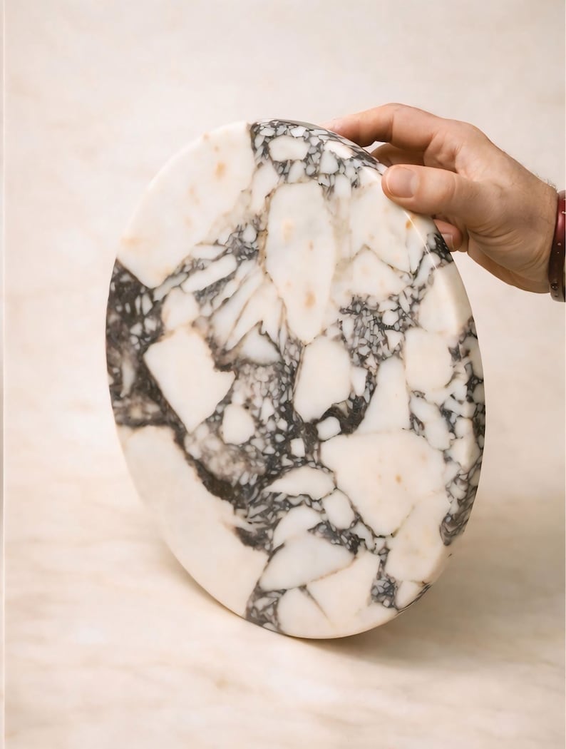 May include: An oval marble chopping board with a distinctive pattern of white, black, and tan veining. The board is held by a hand, highlighting its polished surface and natural stone variations. A decorative kitchen item.