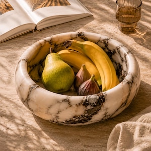 May include: A marble fruit bowl with a pear, bananas, and figs. The bowl has a white and gray marbled pattern. A glass of water and an open book are in the background.