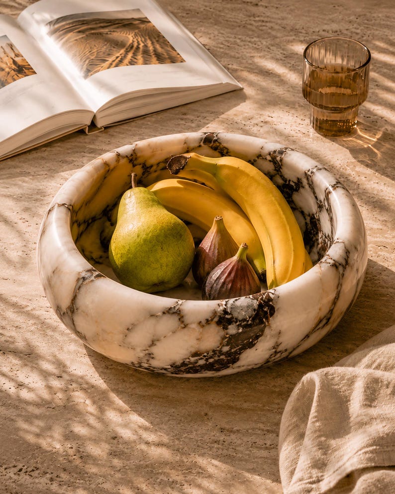 May include: A marble fruit bowl with a pear, bananas, and figs. The bowl has a white and gray marbled pattern. A glass of water and an open book are in the background.