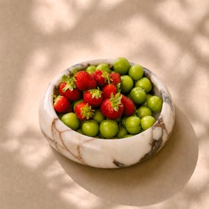 May include: A marble bowl filled with fresh strawberries and green plums. The bowl has a white base with brown veining. The strawberries are bright red with green stems, and the plums are a vibrant green color.