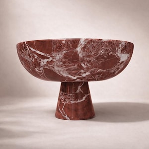 Op de afbeelding: Een rode marmeren kom met witte adering, op een bijpassende voet. De kom heeft een breed, ondiep ontwerp, ideaal voor het presenteren van fruit of decoratieve items. De natuursteen voegt een vleugje elegantie toe.