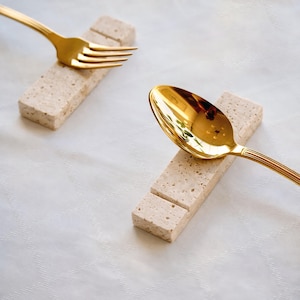 Puede incluir: Un tenedor y una cuchara dorados descansan sobre soportes rectangulares de piedra beige. Los utensilios son de metal dorado brillante. El fondo es un mantel blanco con un patrón sutil. Accesorios de mesa elegantes.