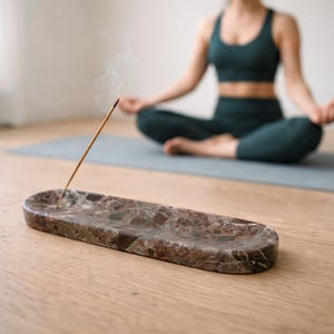 Könnte beinhalten: Ein ovaler Räucherstäbchenhalter aus braunem Marmor mit einem brennenden Räucherstäbchen, das Rauch abgibt. Der Halter steht auf einem Holzboden. Im Hintergrund befindet sich eine Person in einer Yoga-Position, die dunkle grüne Sportkleidung trägt.