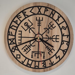 Könnte beinhalten: Eine runde Wanduhr mit hellbrauner Holzmaserung. Das Zifferblatt zeigt in der Mitte ein schwarzes Vegvisir-Symbol, umgeben von einem Ring aus schwarzen Runenzeichen. Sie hat zwei schwarze Zeiger für Stunden und Minuten sowie einen roten Sekundenzeiger.