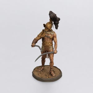 May include: A detailed miniature figurine of a gladiator standing on a circular base. The figure wears a gold-toned helmet with a dark plume, a shoulder guard, and a loincloth. It holds two curved silver-colored blades in a combat stance.