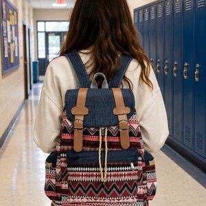 May include: A backpack featuring a dark blue denim top flap and base, accented with tan faux leather straps and silver buckles. The main body displays a geometric pattern in shades of red, black, and white, with a drawstring closure and side pockets.