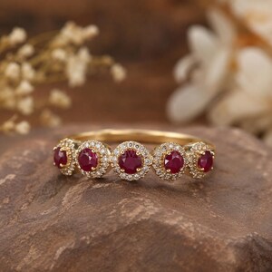 May include: A gold ring featuring five round, deep red rubies arranged in a row. Each ruby is surrounded by a halo of small, clear white diamonds. The band is crafted from polished yellow gold, showcasing a classic design with multiple stones.