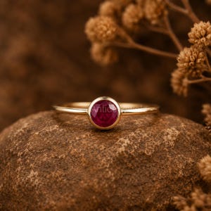 Op de afbeelding: Een gouden ring met een ronde, gefacetteerde, dieprode robijn in een gladde zetting. De ringband is dun en gepolijst, met een strak ontwerp dat de levendige kleur en helderheid van de centrale edelsteen op een duidelijke manier benadrukt.