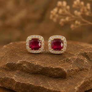 May include: A pair of gold stud earrings featuring cushion-cut deep red gemstones at the center, each surrounded by a halo of small, clear, sparkling white stones set in a square frame with rounded edges and a polished gold metal finish.
