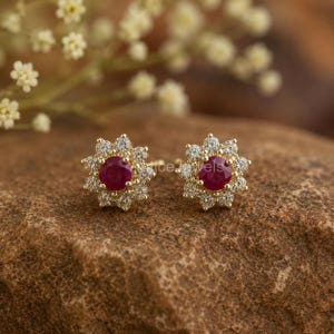 May include: A pair of gold stud earrings, each featuring a round, deep red ruby center stone surrounded by a halo of small, clear, sparkling white stones arranged in a floral pattern, creating a bright and contrasting jewelry design.