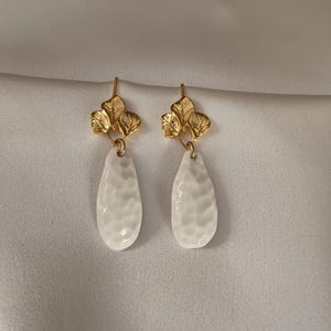 Può includere: Un paio di orecchini pendenti con una parte superiore in metallo dorato a forma di grappolo di tre foglie. Da ogni componente a foglia pende un ciondolo bianco a forma di goccia con una superficie martellata e testurizzata e un gancio.