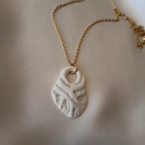 Peut inclure: Un collier avec un pendentif blanc et une chaîne dorée. Le pendentif a une forme irrégulière avec un motif texturé et sculpté. Le collier est présenté sur une surface de couleur neutre.