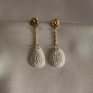 Peut inclure: Une paire de boucles d'oreilles pendantes composées de clous dorés à la texture martelée, reliés par de courtes chaînes dorées à des pendentifs blancs en forme de goutte, dotés d'une surface poreuse rappelant le corail.