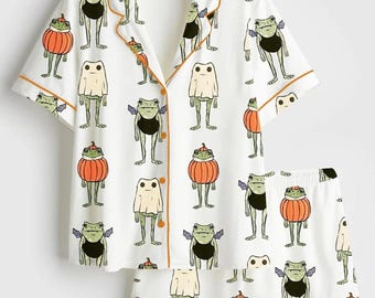 May include: A white short-sleeve button-down pajama shirt and matching shorts set featuring an all-over print of green frogs dressed in various Halloween costumes, including pumpkins, ghost sheets, and bat wings, finished with orange piping trim.