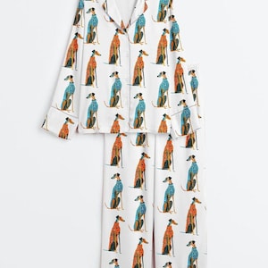 May include: A white pajama set consisting of a long-sleeved button-down shirt with a notched collar and matching wide-leg trousers. The fabric features a repeating pattern of greyhound dogs wearing either orange or blue patterned sweaters.