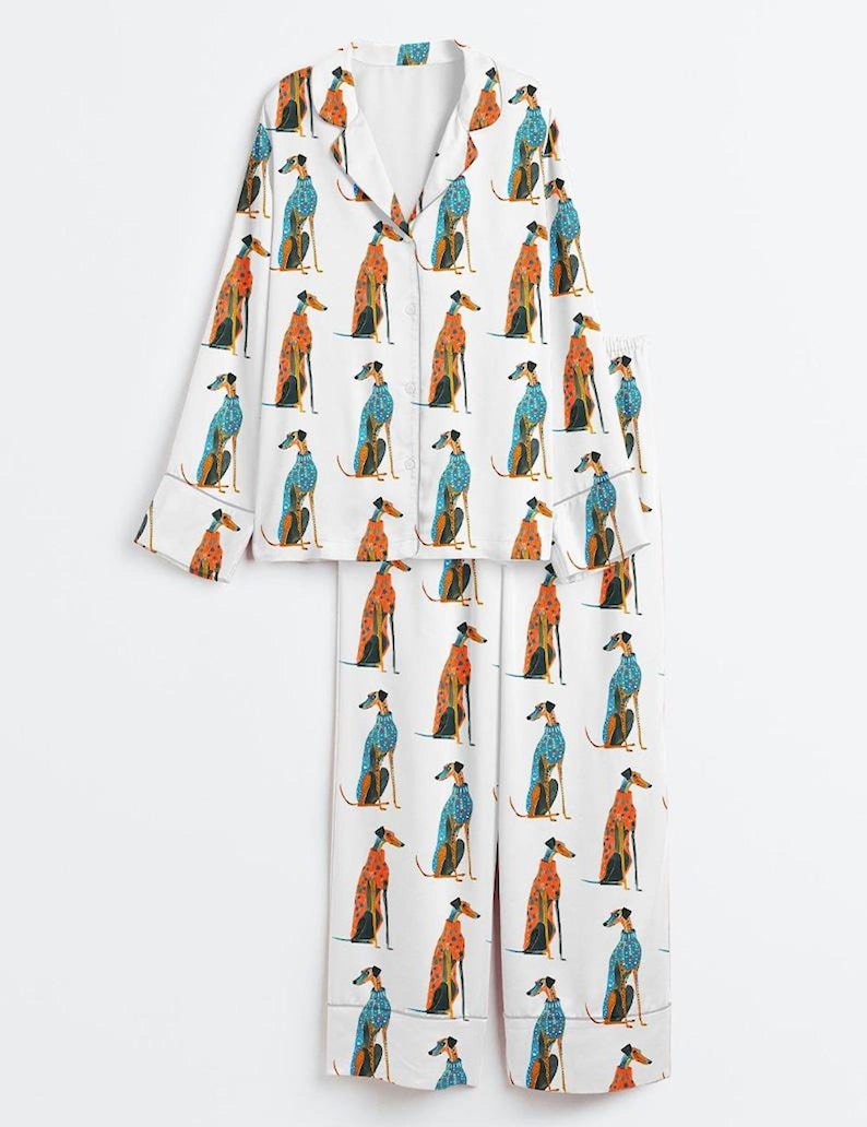 May include: A white pajama set consisting of a long-sleeved button-down shirt with a notched collar and matching wide-leg trousers. The fabric features a repeating pattern of greyhound dogs wearing either orange or blue patterned sweaters.