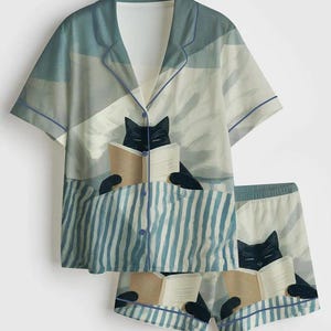 May include: A two-piece pajama set featuring a whimsical design. The top is a button-down shirt with short sleeves, and the shorts have an elastic waistband. Both pieces are adorned with a repeating pattern of a black cat reading a book, set against a blue and white striped background.