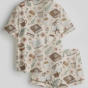 May include: A two-piece pajama set featuring a cream-colored background with a repeating pattern of books, coffee cups, eyeglasses, and flowers in shades of brown, beige, and green. The set includes a short-sleeved button-up shirt and matching shorts.