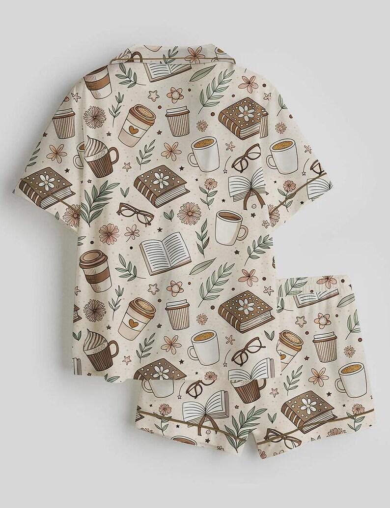 May include: A two-piece pajama set featuring a cream-colored background with a repeating pattern of books, coffee cups, eyeglasses, and flowers in shades of brown, beige, and green. The set includes a short-sleeved button-up shirt and matching shorts.