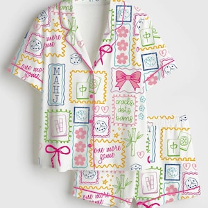 May include: White pajama set with a colorful print featuring dice, flowers, bamboo, and the text "one more game." The set includes a short-sleeved button-up shirt and matching shorts, trimmed with pink piping.