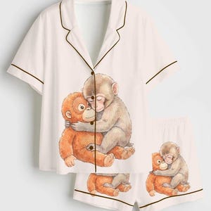 Pu&ograve; includere: Pigiama bianco con top a maniche corte e pantaloncini. Il set presenta un bordo marrone e un'illustrazione ad acquerello di due scimmie che si abbracciano. Top e pantaloncini hanno lo stesso disegno.