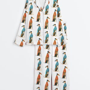 May include: A white pajama set consisting of a long-sleeved button-down shirt with a notched collar and matching wide-leg trousers. The fabric features a repeating pattern of greyhound dogs wearing either orange or blue patterned sweaters.
