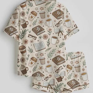 May include: A two-piece pajama set featuring a cream-colored background with a repeating pattern of books, coffee cups, eyeglasses, and flowers in shades of brown, beige, and green. The set includes a short-sleeved button-up shirt and matching shorts.