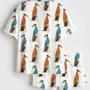 May include: A white short-sleeved button-down shirt and matching shorts set featuring an all-over print of greyhound dogs wearing patterned sweaters in shades of orange, blue, and black. The shirt has a collar and the shorts have an elastic waistband.