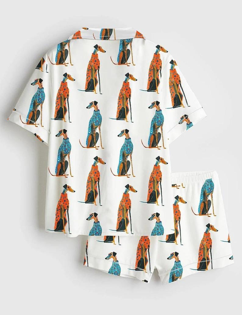 May include: A white short-sleeved button-down shirt and matching shorts set featuring an all-over print of greyhound dogs wearing patterned sweaters in shades of orange, blue, and black. The shirt has a collar and the shorts have an elastic waistband.